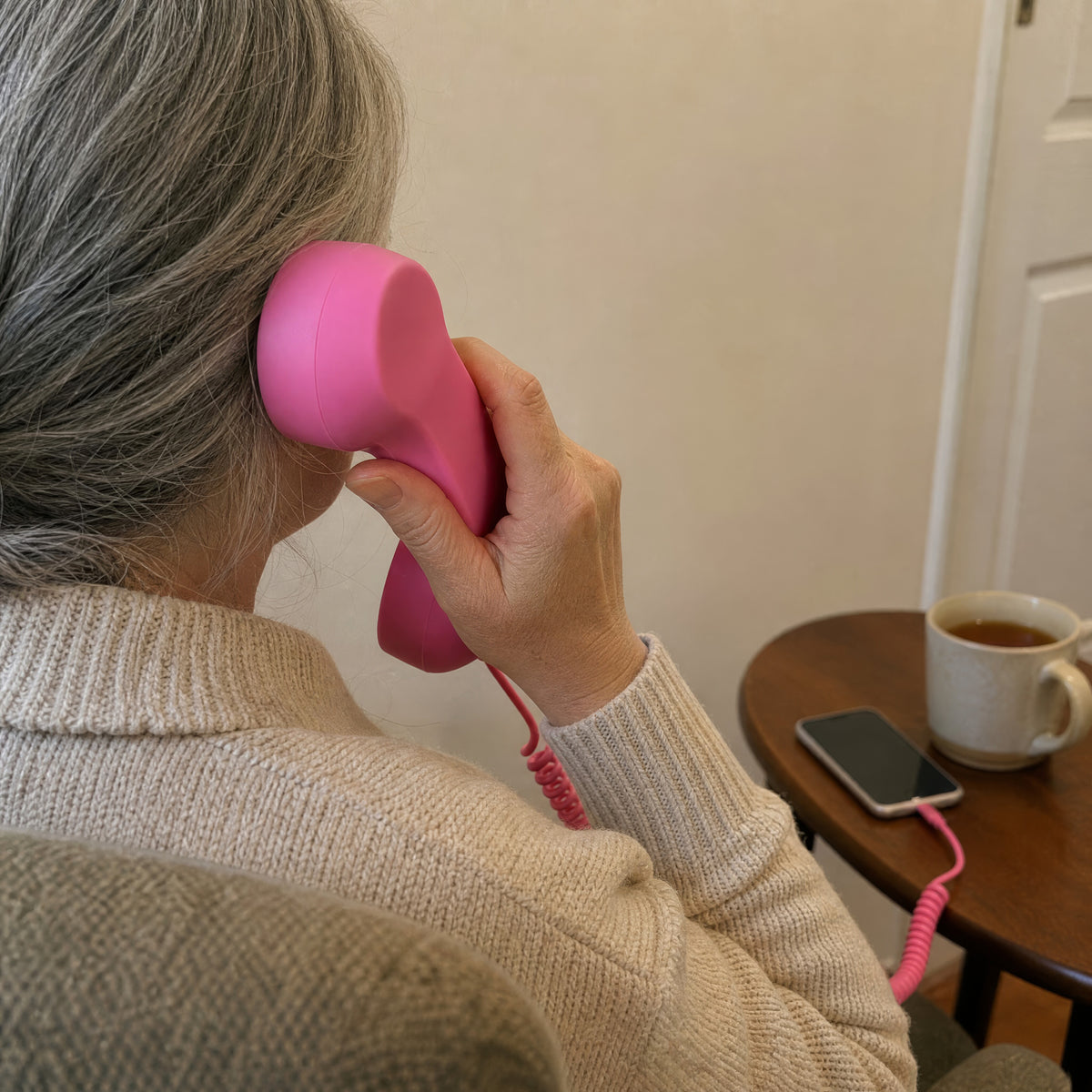 A woman in an armchair holds the pink DialBop handset to her ear, cord running to her phone on the side table