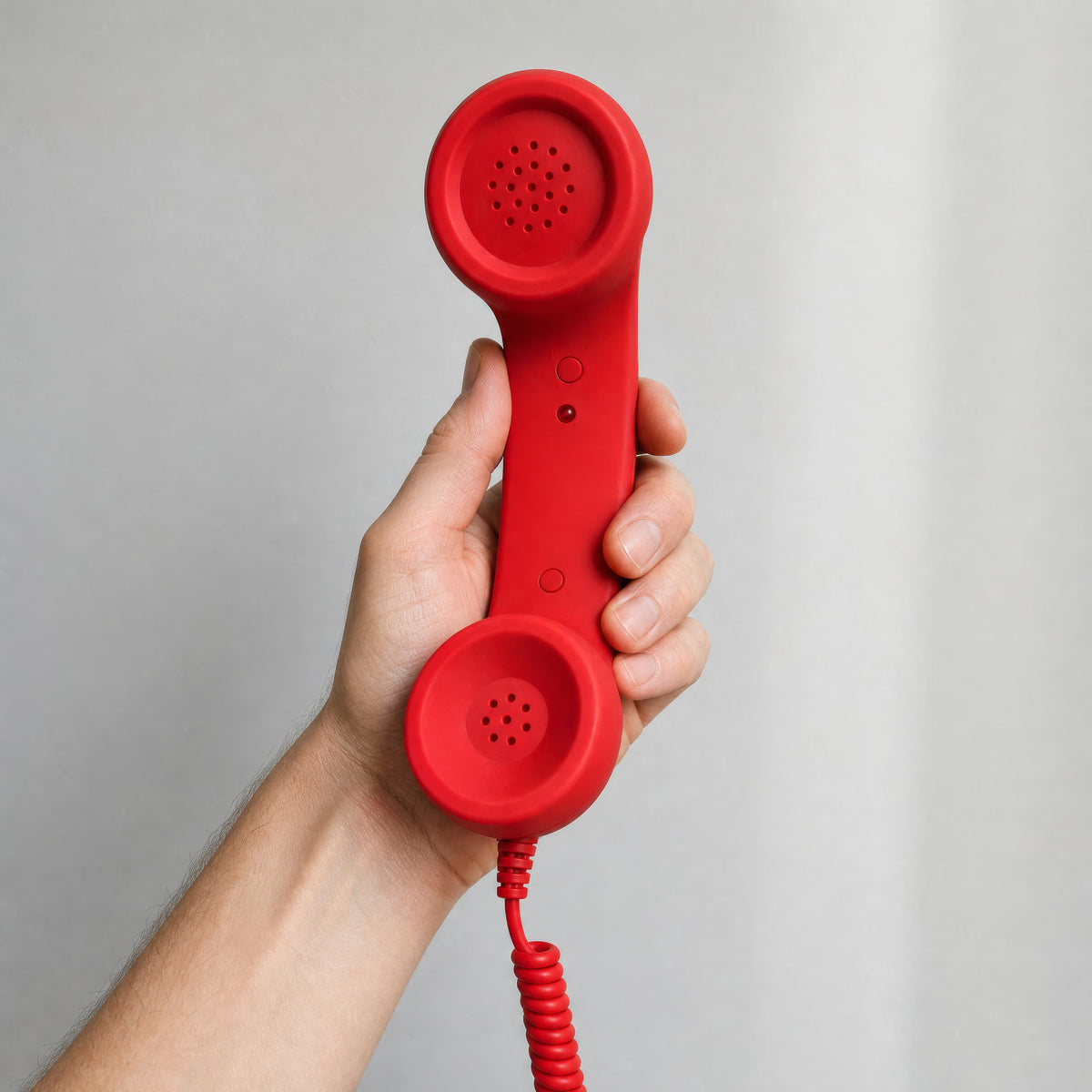 The red DialBop Retro Handset held in one hand against a grey wall, for scale, button and LED facing the camera